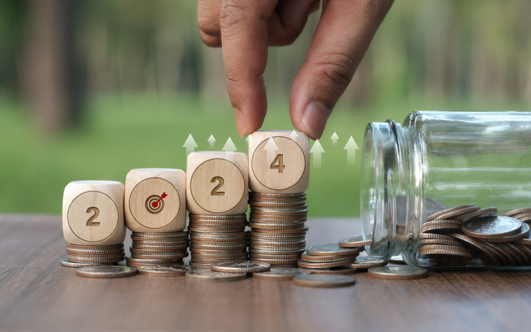 Stacks of coins with wooden blocks showing 2024 and a hand placing a block.